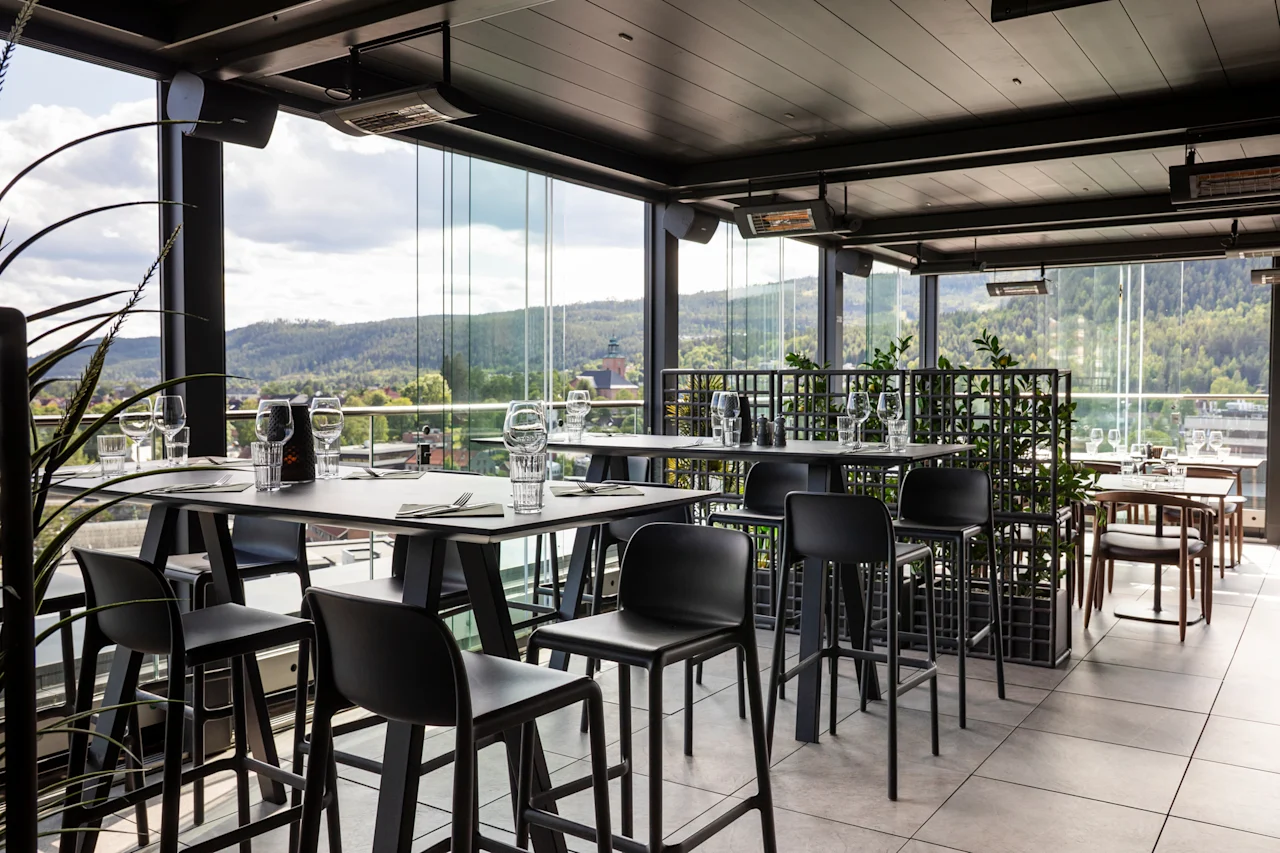 En moderne takterrasse-restaurant med høye bord og barkrakker, dekket for bespisning, med naturskjønn utsikt over grønne åser.