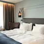 Et koselig hotellrom med en seng med hvitt sengetøy og et blått pledd, en lenestol og varm belysning.
