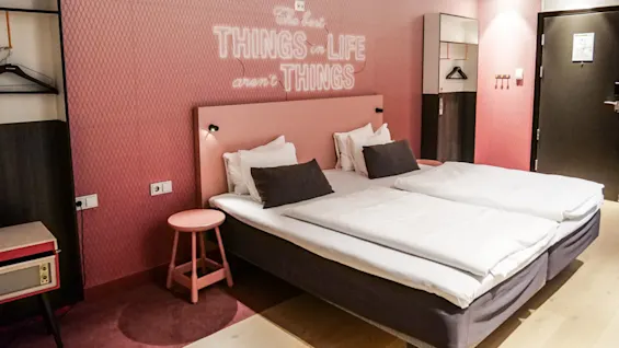Et hotellrom med to senger, hvitt sengetøy og svarte og rosa puter; rosa vegger viser teksten "The best THINGS in LIFE aren’t THINGS" samt garderobe og dør.