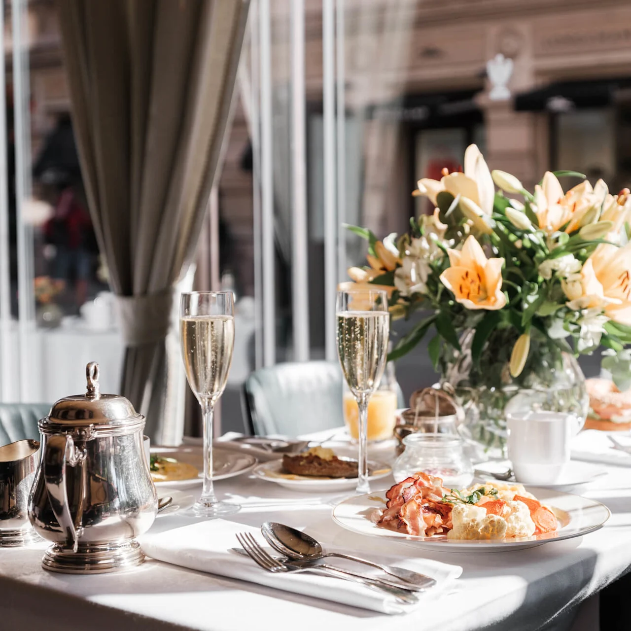 A well-set breakfast table with champagne, silverware, and a colorful flower arrangement in a sunny, windowed room at Hotel Kämp in Helsinki.