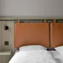 Brunt vadderat huvudgavel fäst vid en vägg stöder två vita kuddar på en säng i ett minimalistiskt sovrum. En liten hylla med en lampa och ett eluttag är intill.