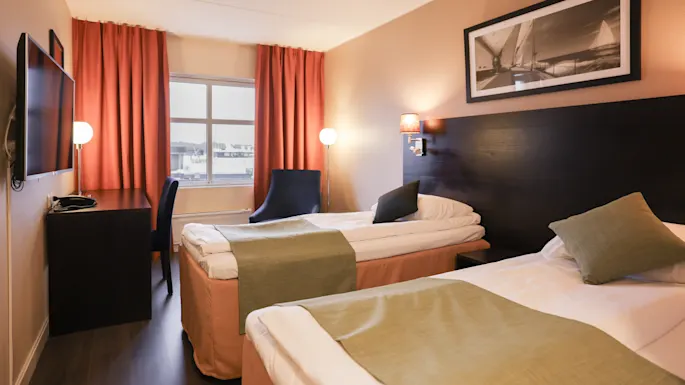 Two neatly made twin beds with green throws are positioned in a hotel room featuring a desk, chair, and a window with orange curtains. A black-and-white sailing photo adorns the wall.