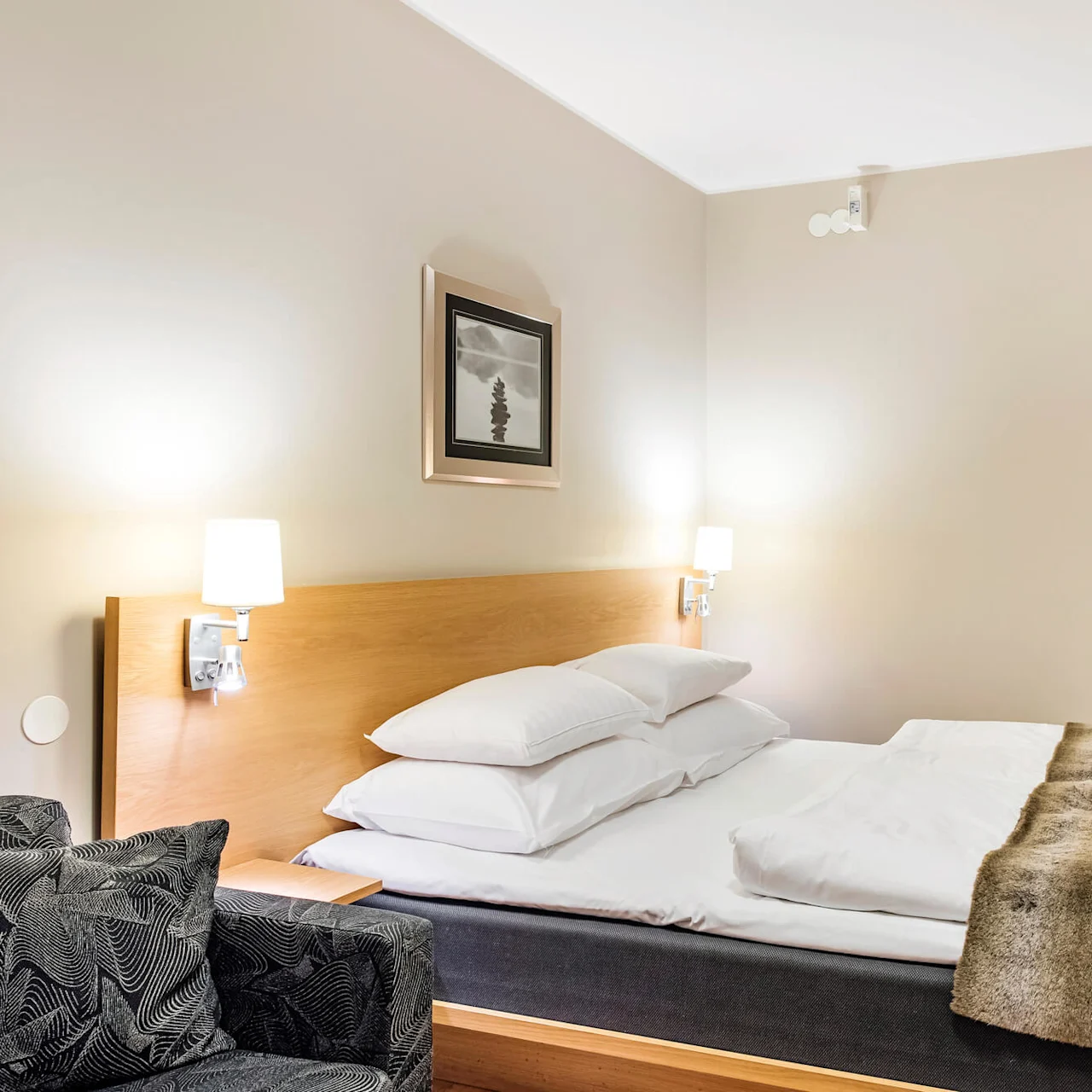 En pent oppredd seng er sentrert i et hotellrom, flankert av to vegglamper. Et abstrakt maleri henger over trehodegjerdet, og en lenestol står like ved.