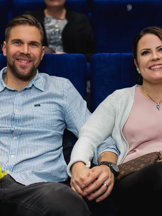 Hymyilevä pariskunta pitelee toisiaan kädestä elokuvateatterissa tai auditoriossa.
