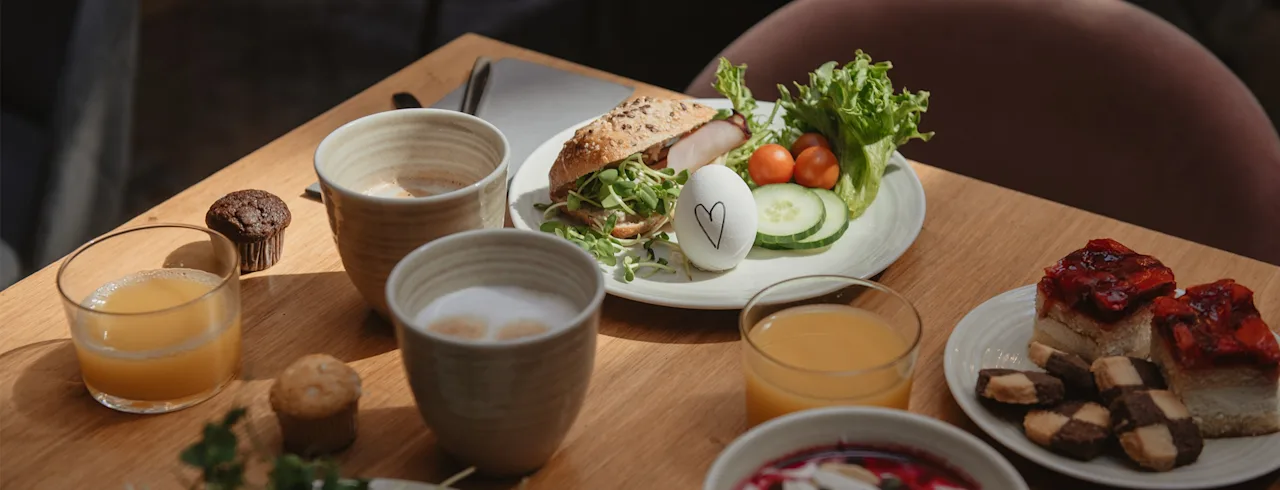 En mysig frukostscen med en mängd olika maträtter och drycker på ett träbord, inklusive en smörgås, ett ägg med ett hjärta, bakverk och kaffe.