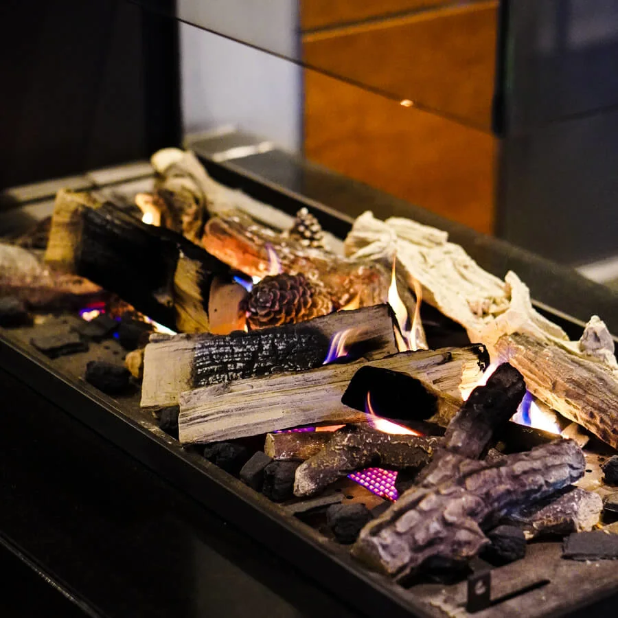 Logs lie atop glowing embers in an indoor electric fireplace, surrounded by reflective glass, creating a warm, cozy ambiance.