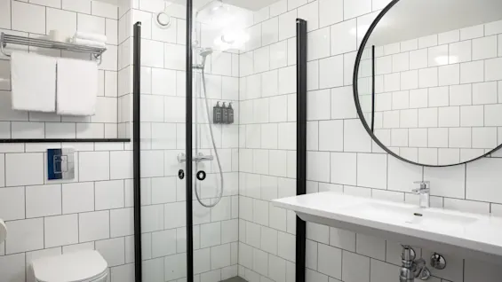 Et moderne badeværelse har en brusekabine med glas, væghængte toiletartikler, et stort rundt spejl over en hvid vask, hvide flisebelagte vægge og et pænt arrangeret håndklædestativ.