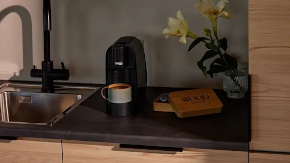 A coffee maker with a mug sits on a dark kitchen counter. Nearby are lilies in a vase, and a wooden box labeled "WOOD Coffee Beans." Light wood cabinets surround.