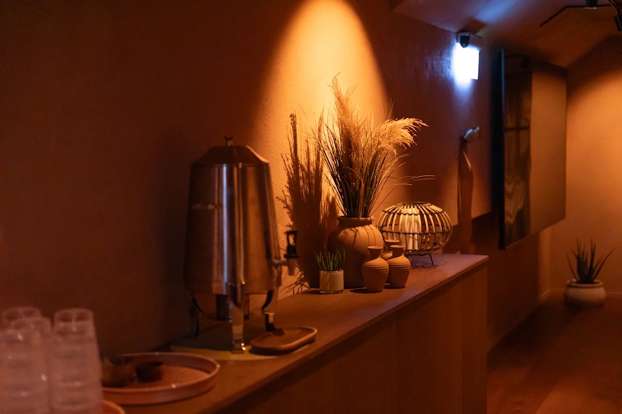 Viihtyisä nurkkaus, jossa on juoma-automaatti, koristekasveja ja kuppeja tiskillä, valaistuna lämpimällä valolla.