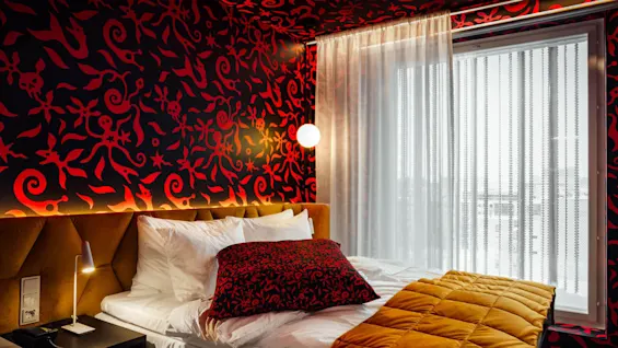 Bed with white sheets and a red-patterned pillow is situated in a room with vibrant, swirling red and black wallpaper. A window with sheer curtains illuminates the space.