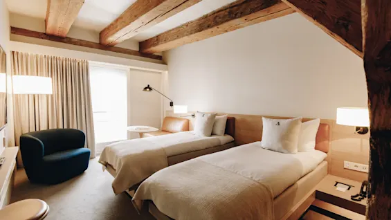 Ett mysigt hotellrum med två enkelsängar, synliga takbjälkar i trä och en bekväm fåtölj vid fönstret.