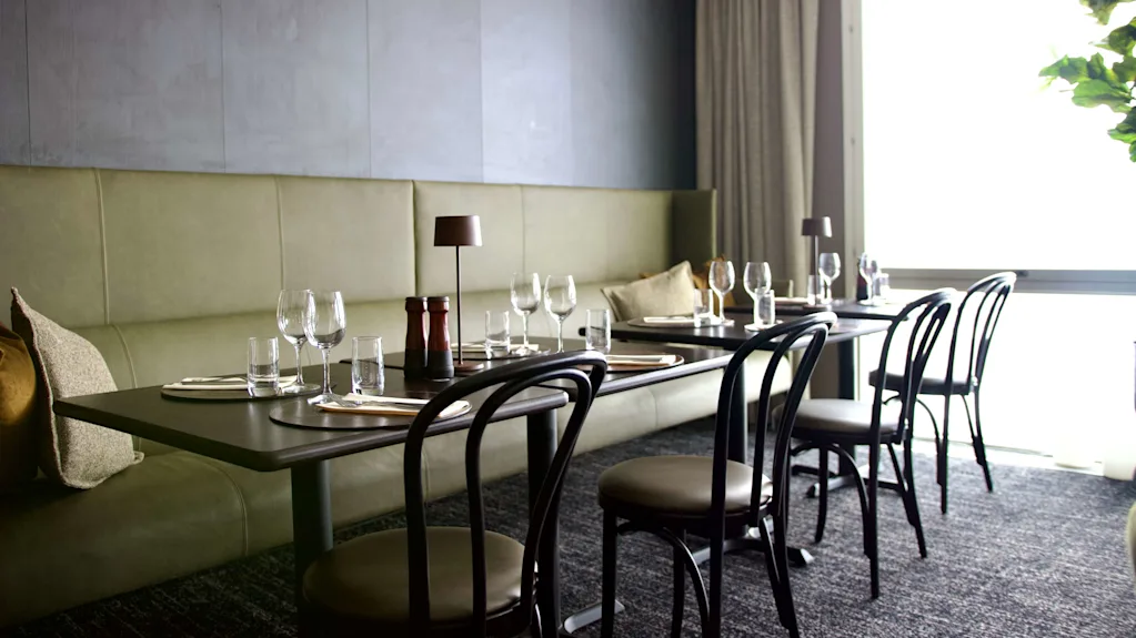 Ett restaurangbord dukat med vinglas och bestick, placerat på ett mörkt golv nära en ljusgrön vadderad sittplats, under mjuk belysning i ett modernt matområde.
