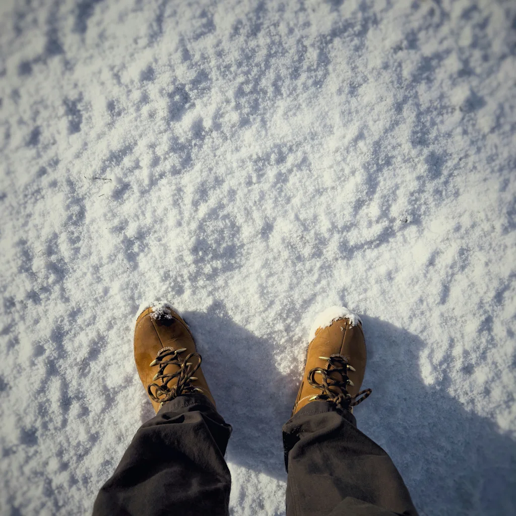 En persons fødder i brune støvler, der står i frisk sne, med sollys, der kaster skygger.