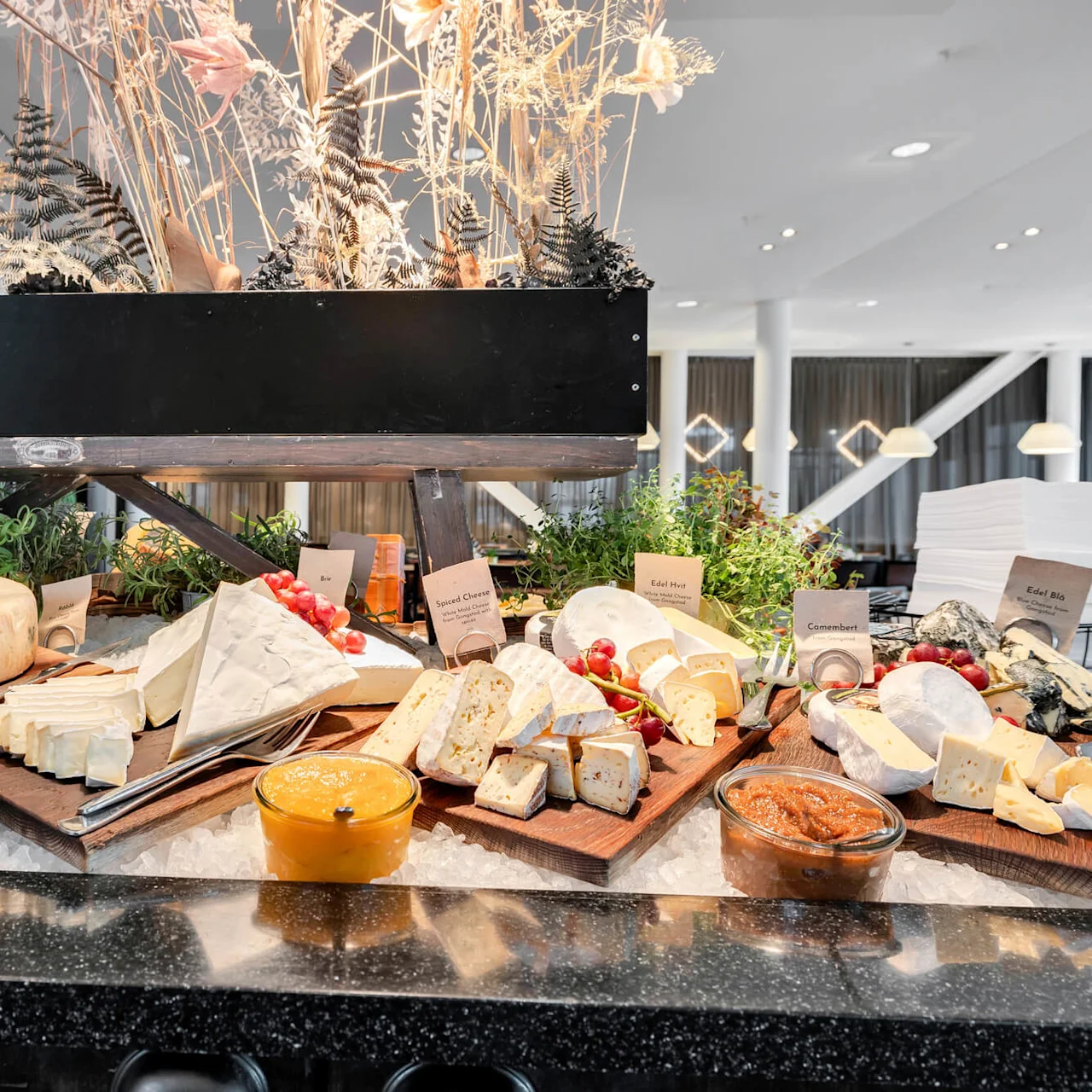 Cheeses are displayed on wooden boards with labels, surrounded by fig marmalade and herbs, in a well-lit buffet setting. Labels include "Brie", "Spiced Cheese", "Edil Basics", "Edil Hot", "Camembert", "Edil Bis", "Fig Marmalade".