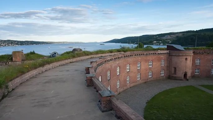 A curved brick fort overlooks a serene body of water, surrounded by grassy hills and distant forests under a blue sky with fluffy clouds.