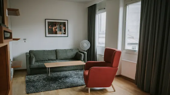 En rød lænestol vender mod en grøn sofa i et lyst rum med trægulv, sofabord og et indrammet fotografi på væggen. Store vinduer lader naturligt lys komme ind.