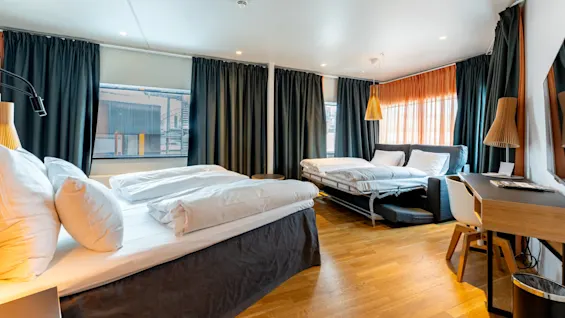 Et hotelværelse indeholder to pænt redte senge med hvidt sengetøj; naturligt lys filtrerer gennem store vinduer med mørke gardiner. Et skrivebord og siddeområde tilføjer funktionalitet.