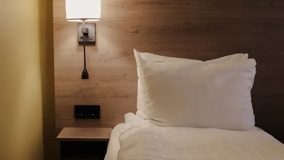 A neatly made bed with white linens and green blanket, next to a wall-mounted lamp, in a cozy, wood-panelled bedroom.