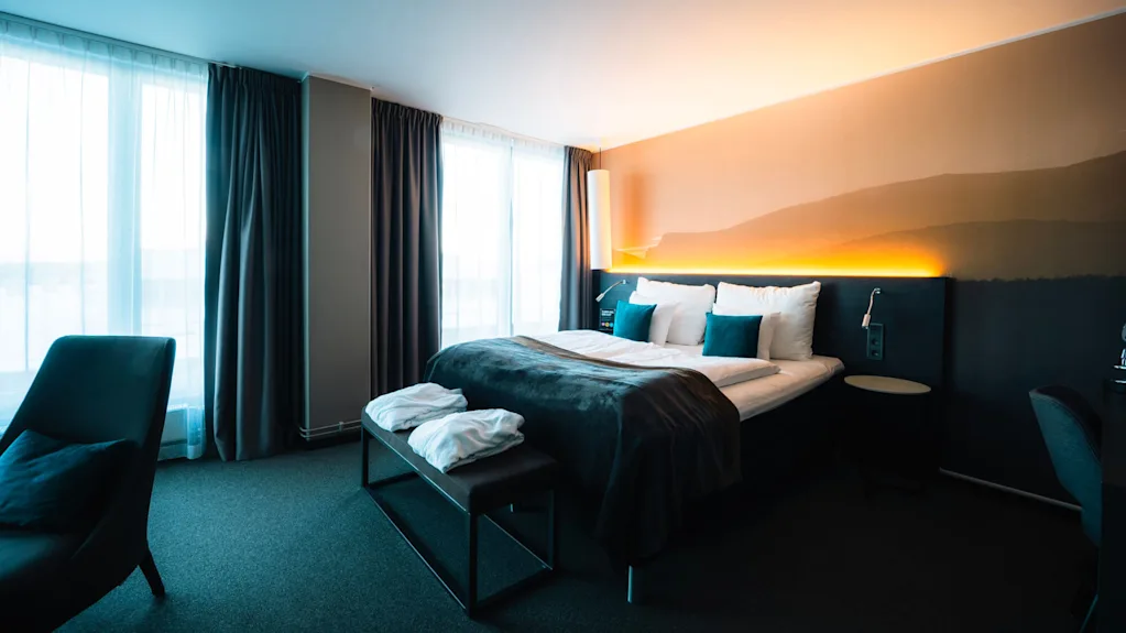En snyggt bäddad säng med vita och teal kuddar, täckt av en mörk filt, står bredvid stora fönster med skira gardiner, vilket skapar en ljus och inbjudande hotellrumsmiljö.
