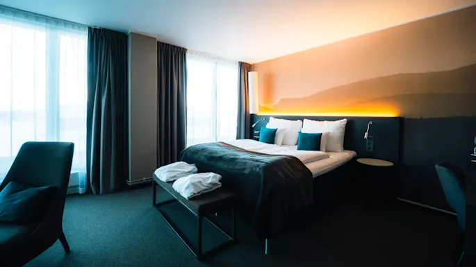 En pent oppredd seng med hvite og blågrønne puter, dekket av et mørkt teppe, står ved siden av store vinduer med gjennomsiktige gardiner, som skaper en lys og innbydende hotellromatmosfære.