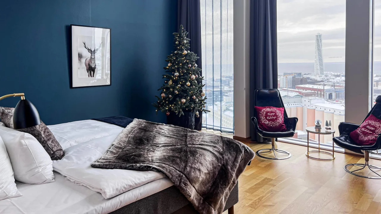 A bedroom with a Christmas tree and city view, dark blue walls, snowflake decorations, and two swivel chairs near a window.