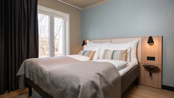 Ett mysigt hotellrum med en bekväm säng, randiga kuddar och ett fönster med mörka gardiner, inbjudande till avkoppling.