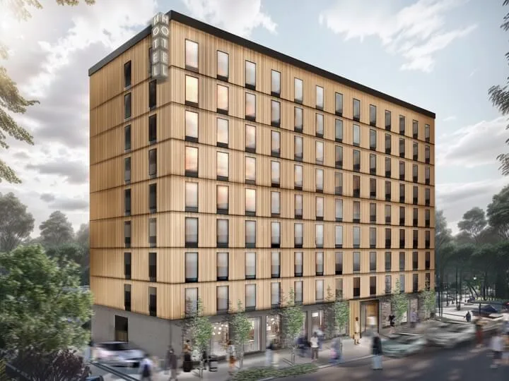 Rakennus seisoo korkeana puujulkisivulla, merkintä "HOTEL." Ihmisiä ja autoja liikkuu puiden reunustamaa katua pitkin. Taivas on pilvinen pehmeän valon siivilöityessä läpi.