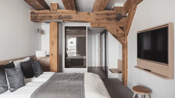 A bed with gray bedding is positioned under wooden beams in a modern room, facing a wall-mounted TV. A mirror reflects a windowed view.