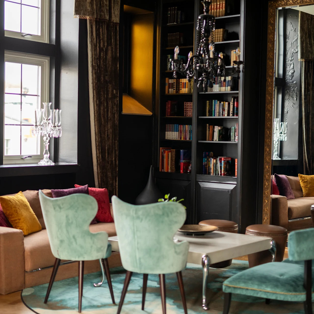 A cozy, elegant seating area features turquoise chairs around a table, surrounded by bookshelves and a brown sofa with colorful cushions, in a room with large windows and a decorative chandelier.