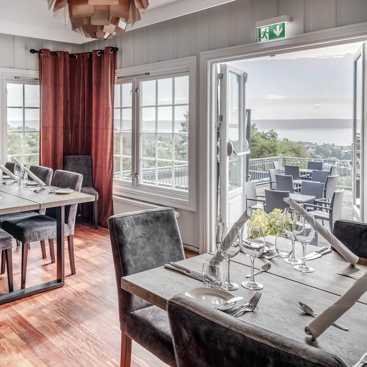 Et spiserom med bord dekket med glass og servietter, med utsikt over en naturskjønn uteservering gjennom åpne balkongdører.