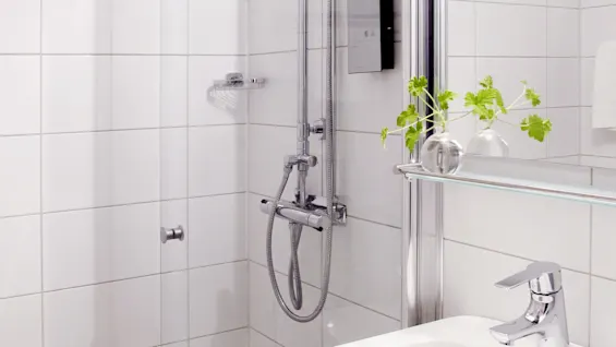 Dusj med doble hoder er festet i et moderne, hvitfliset bad. En liten servant og en plante i en glassvase er i nærheten, noe som forsterker det rene, minimalistiske designet.