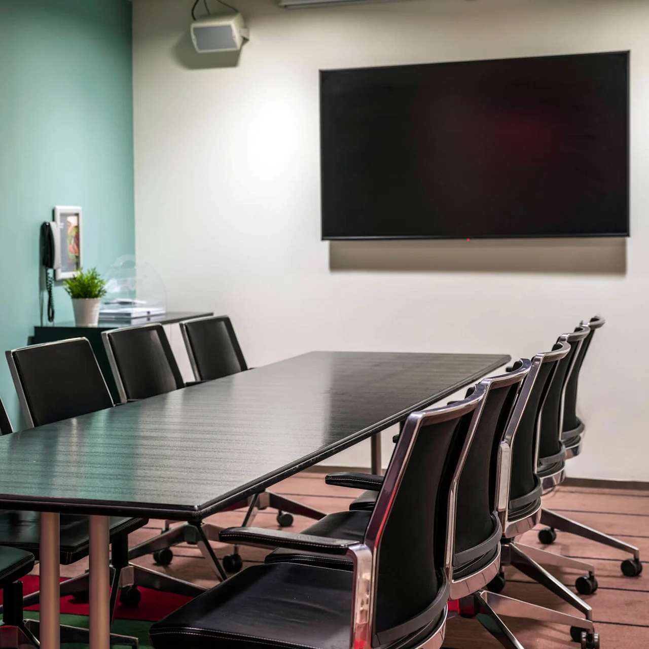 A long conference table surrounded by black chairs is in an office meeting room. A large blank screen is mounted on the wall, and a green plant sits on a side cabinet.