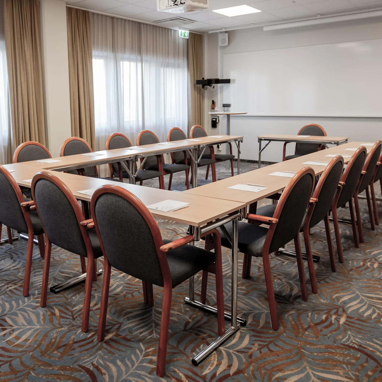 Conference table set in U-shape with chairs, paper pads, and pens; facing a whiteboard in a carpeted room with large windows and curtains.