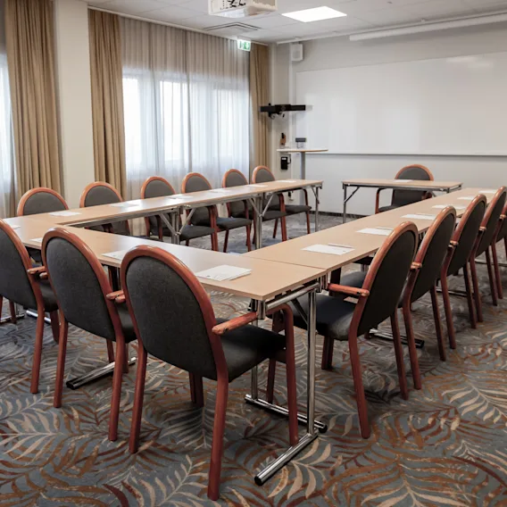 Konferensbord i U-form med stolar, pappersblock och pennor; vetter mot en whiteboard i ett rum med heltäckningsmatta och stora fönster och gardiner.
