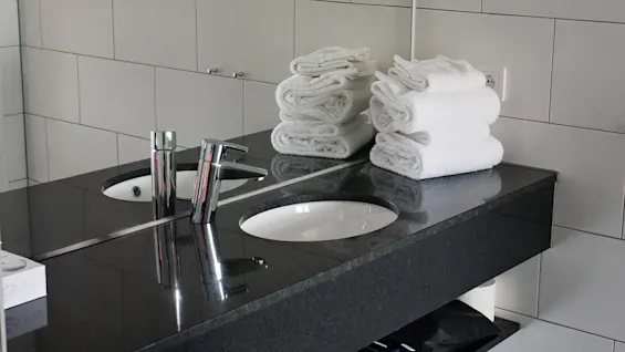 Moderni kylpyhuone, jossa on kaksi pesuallasta, puhtaat valkoiset pyyhkeet ja tyylikäs tumma työtaso, valmiina vieraille.