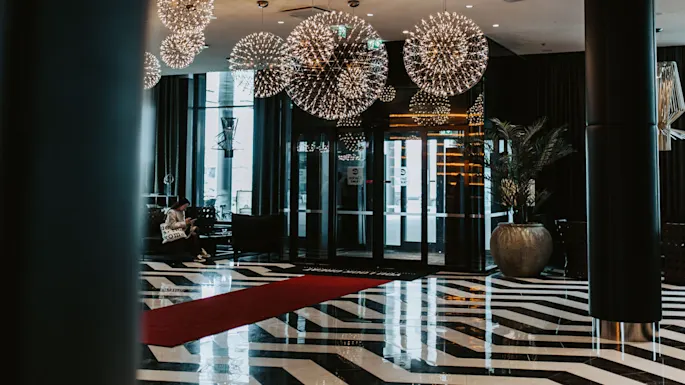Lysekroner henger over et blankt, mønstret gulv; en person sitter og leser i en stilren, moderne lobby med store glassdører, potteplanter og en fremtredende rød løper.