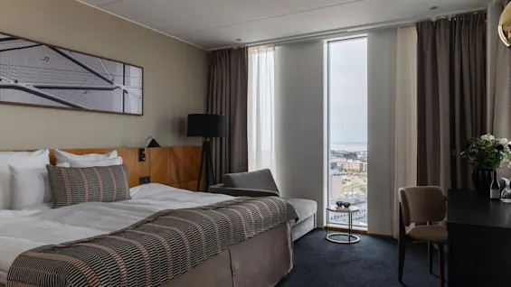 Et moderne hotellrom med en komfortabel seng, et stort vindu med utsikt over byen og et skrivebordsområde.