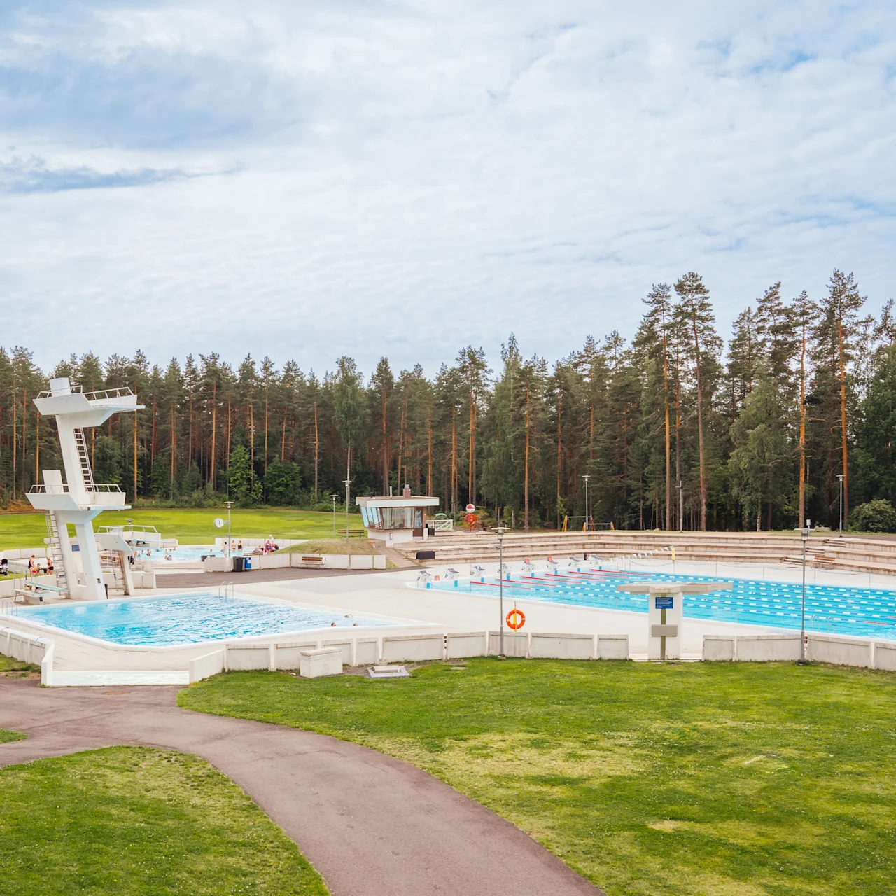 Public swimming pool with diving platforms surrounded by green grass and tall trees under a partly cloudy sky. Pathways lead to the pool area. No text is present.