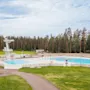 Yleisöuimala hyppytorneilla, jota ympäröi vihreä ruoho ja korkeat puut osittain pilvisellä taivaalla. Polut johtavat allasalueelle. Tekstiä ei ole.