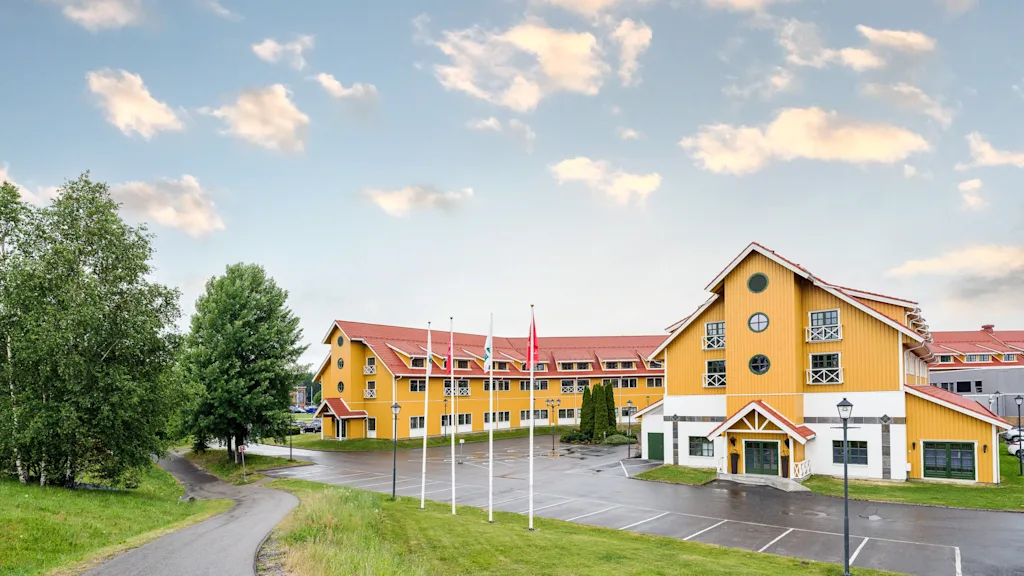 En stor, gul byggnad med röda tak står bredvid en parkeringsplats, omgiven av träd och en gångväg under en delvis molnig himmel. Fem flaggstänger står framför.
