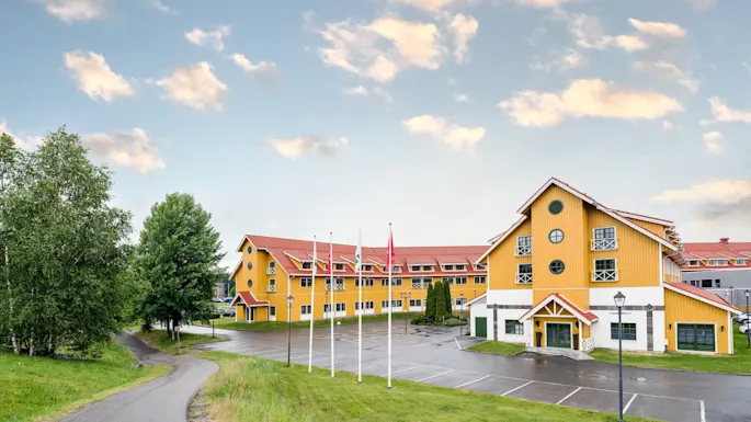A large, yellow building with red roofs stands next to a parking lot, surrounded by trees and a pathway under a partly cloudy sky. Five flagpoles line the front.