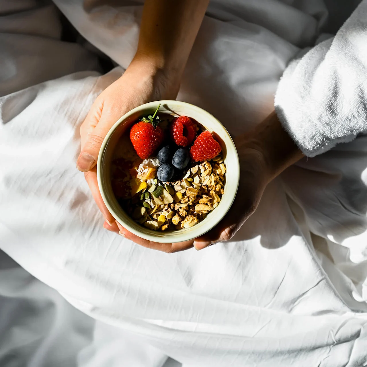 En skål med müsli og bær holdes af to hænder. Personen har en hvid morgenkåbe og sidder på hvidt sengetøj, med sollys der kaster bløde skygger.