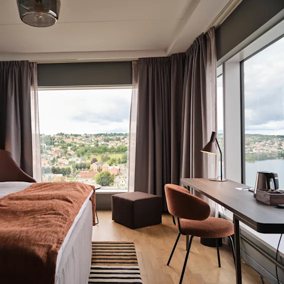 Hotellrom viser en pent oppredd seng, brunt teppe og lenestol. Skrivebord med lampe og vannkoker vender mot store vinduer med byutsikt under spredte skyer.