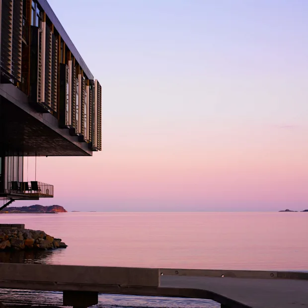 En modern byggnad hänger över lugnt havsvatten i skymningen, mot en pastellhimmel.