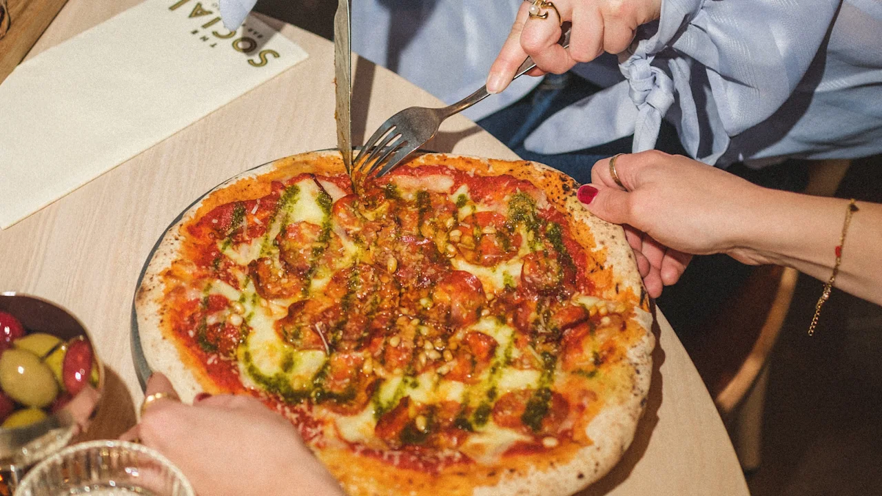 Henkilö leikkaa herkullista pesto- ja pepperonipizzaa, jakaen aterian ystävien kanssa. Servetissä lukee SOCIAL.