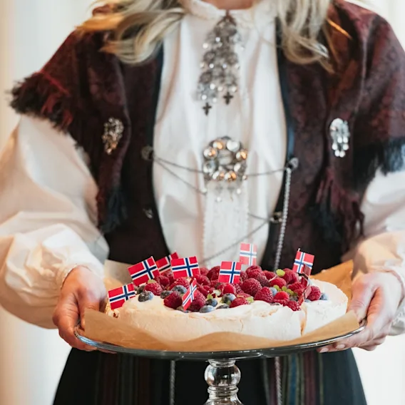 Nærbilde av en person i bunad som holder en pavlova toppet med bringebær, blåbær og små norske flagg.