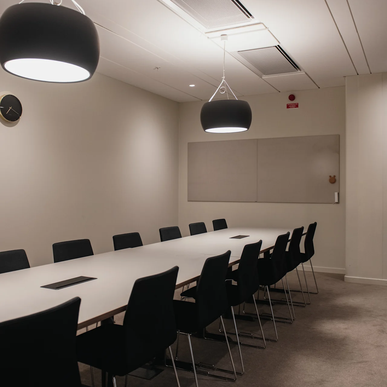 A long conference table surrounded by black chairs sits in a well-lit meeting room with beige walls. Overhead, two large pendant lights hang, and a clock is mounted on the wall.