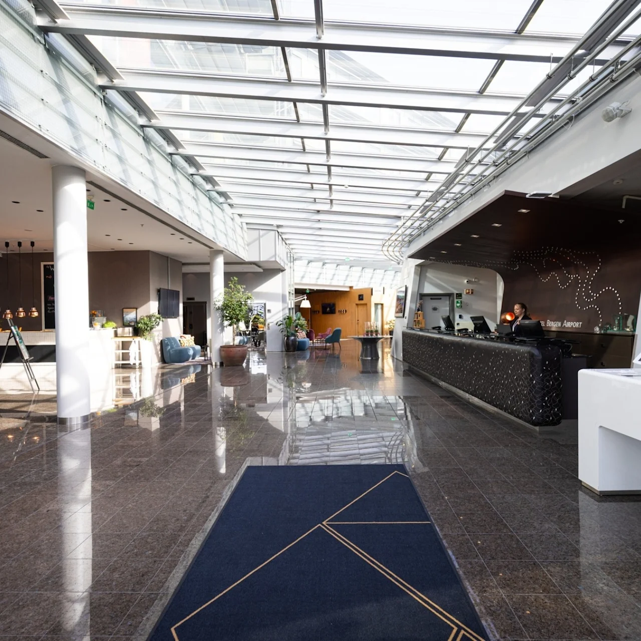 A hotel lobby with a shiny tiled floor. A receptionist stands behind a black counter on the right, welcoming guests. Potted plants and modern furniture adorn the spacious, sunlit corridor.