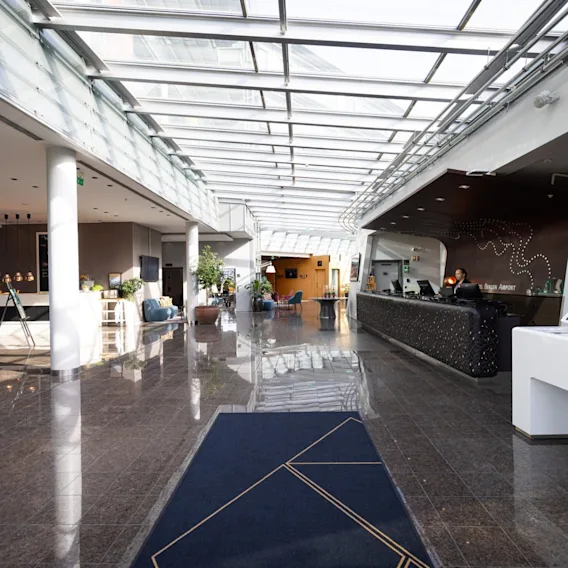 En hotellobby med glinsende fliser. En resepsjonist står bak en svart skranke til høyre og ønsker gjester velkommen. Potteplanter og moderne møbler pryder den romslige, solfylte korridoren.