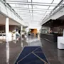 En hotellobby med glinsende fliser. En resepsjonist står bak en svart skranke til høyre og ønsker gjester velkommen. Potteplanter og moderne møbler pryder den romslige, solfylte korridoren.